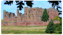 Kenilworth Castle ruins in Warwickshire, England, with towering red sandstone walls surrounded by grassy fields and trees, viewed under a partly cloudy sky.