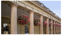 Exterior of the Royal Spa Pump Room, featuring a row of classical stone columns with hanging baskets of pink flowers, and a shop window visible on the left beneath a clear sky.