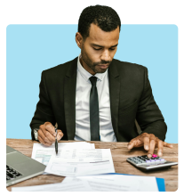 Man in a business suit sitting at a desk, working with documents and using a calculator, with a laptop open beside him.